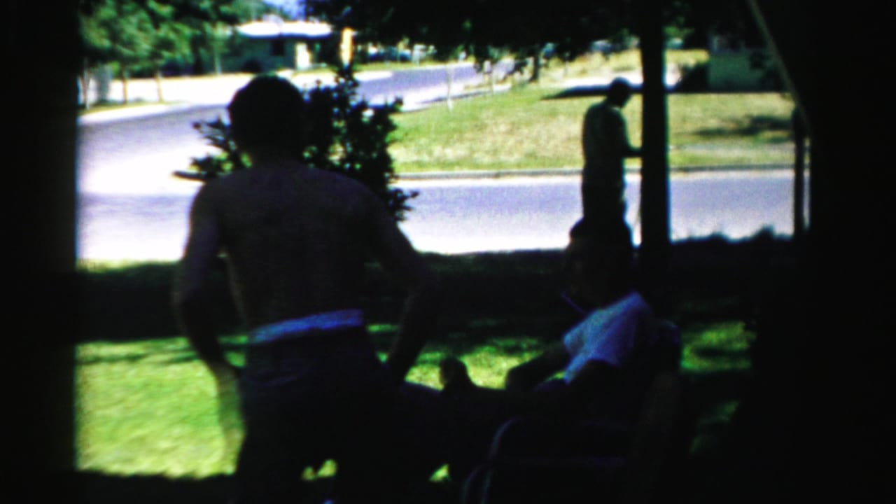 1957 Normal, Illinois home-movie frame showing seated adults in deep shade, a green lawn, and a standing figure by the road.