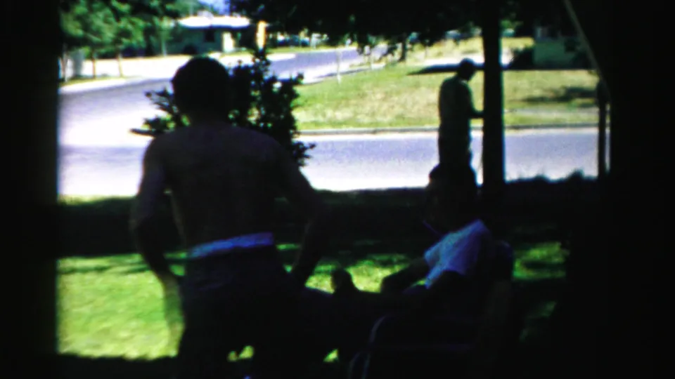 1957 Normal, Illinois home-movie frame showing seated adults in deep shade, a green lawn, and a standing figure by the road.