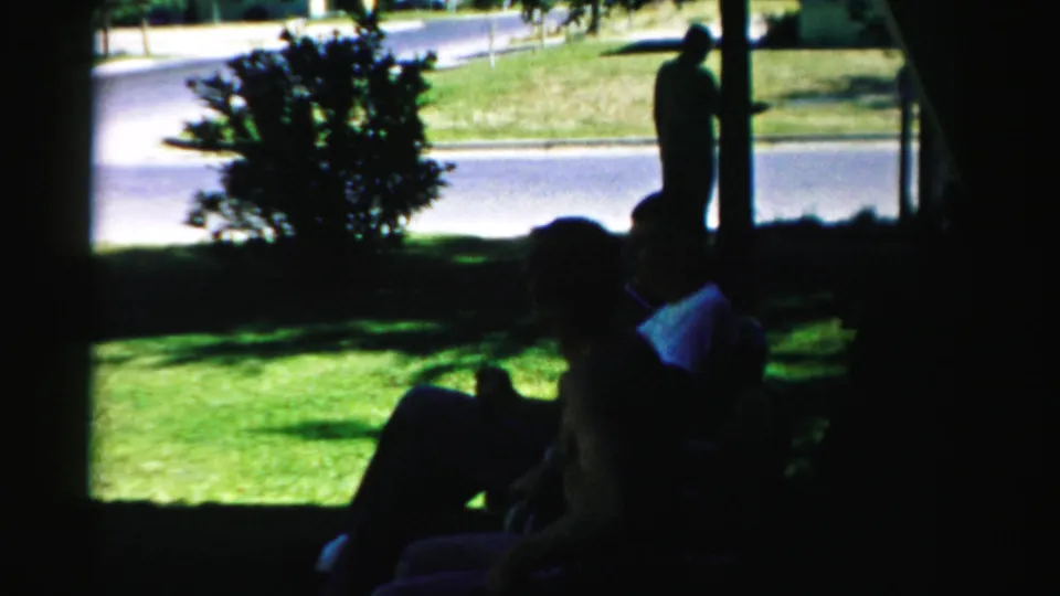 Shaded 1957 Normal, Illinois yard view with a seated adult, tree shadows, lawn, roadside, and a standing figure in sunlight.