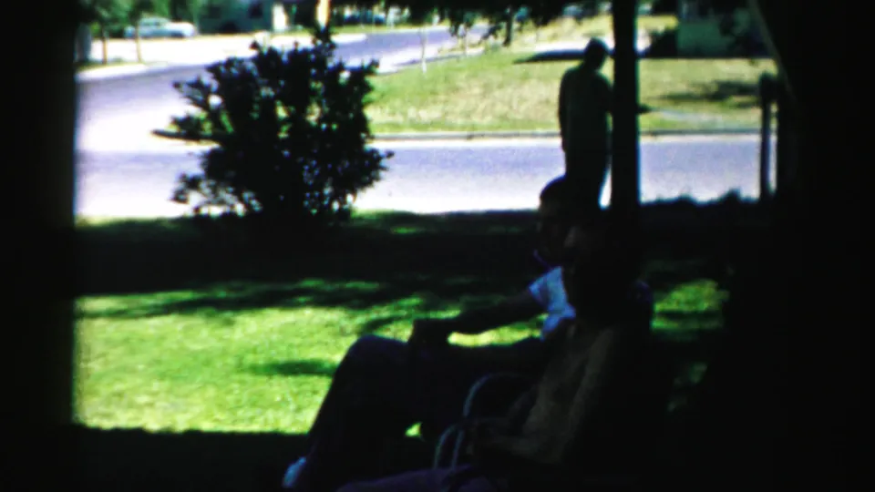 1957 Normal, Illinois frame with seated adults in silhouette, a standing person near the curb, trees, lawn, and residential road.