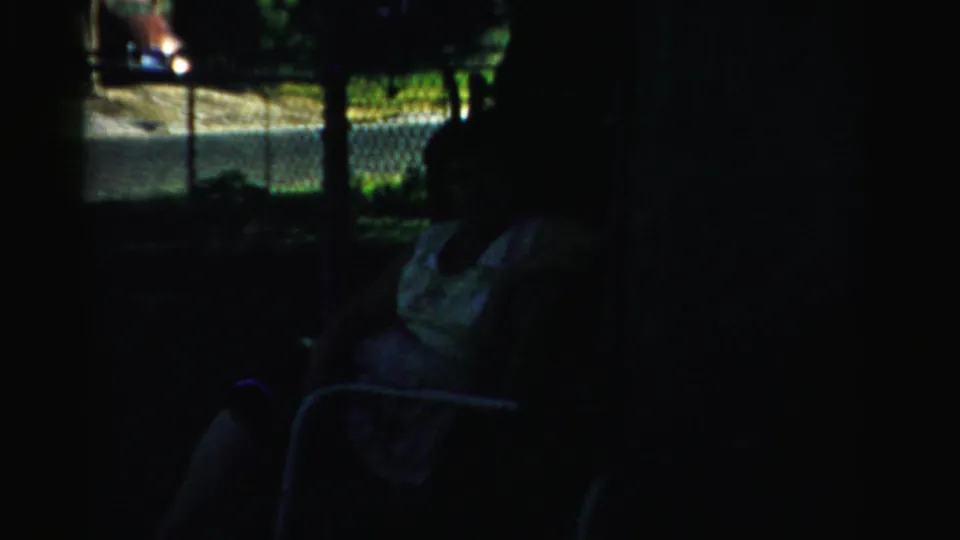 Late frame from the 1957 Normal, Illinois home movie with a seated figure in shade, lawn, fence, road, and bright yard beyond.