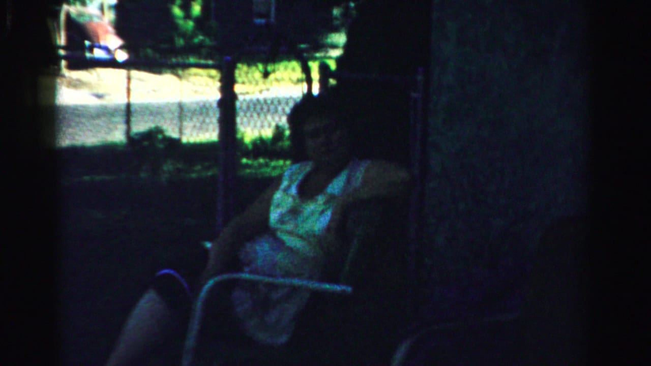 In Normal, Illinois home-movie footage, a woman sits in a metal chair under shade with a chain-link fence and greenery behind her.