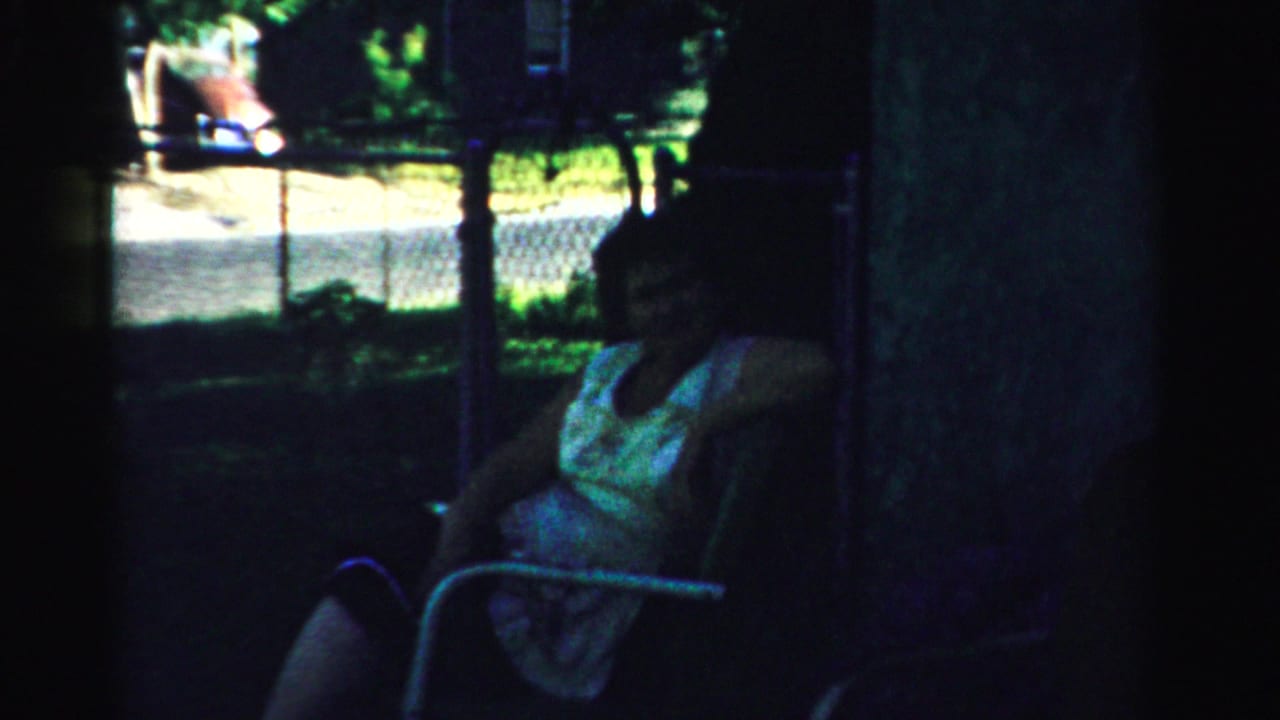 A woman in a light dress appears seated outdoors in shaded 1950s Normal, Illinois footage, with fencing and greenery behind her.