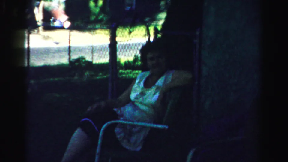 The 8mm frame shows a woman seated in a metal lawn chair in Normal, Illinois, with deep shade and a sunlit fence beyond.