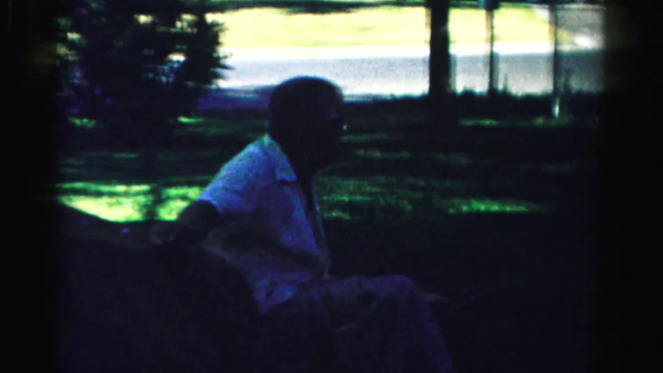 A man in a light shirt sits on a bench or low seat in shaded Normal, Illinois home-movie footage, with trees and roadway beyond.