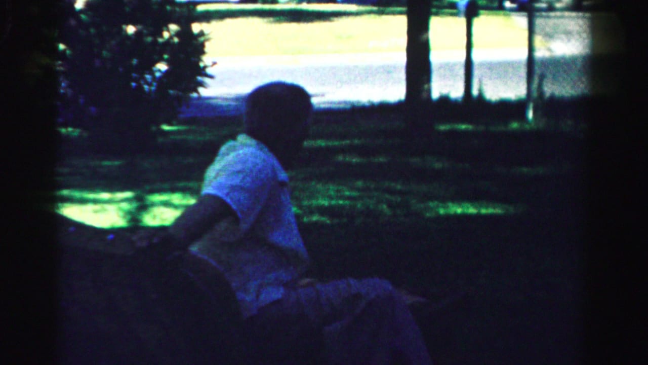 Older man in a light short-sleeve shirt sits outdoors in shadow in Normal, Illinois, 1957 home-movie footage.