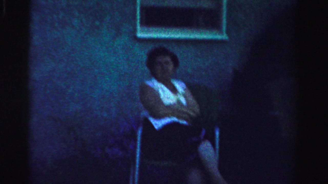 Older woman seated outside against a house wall with a window above her.