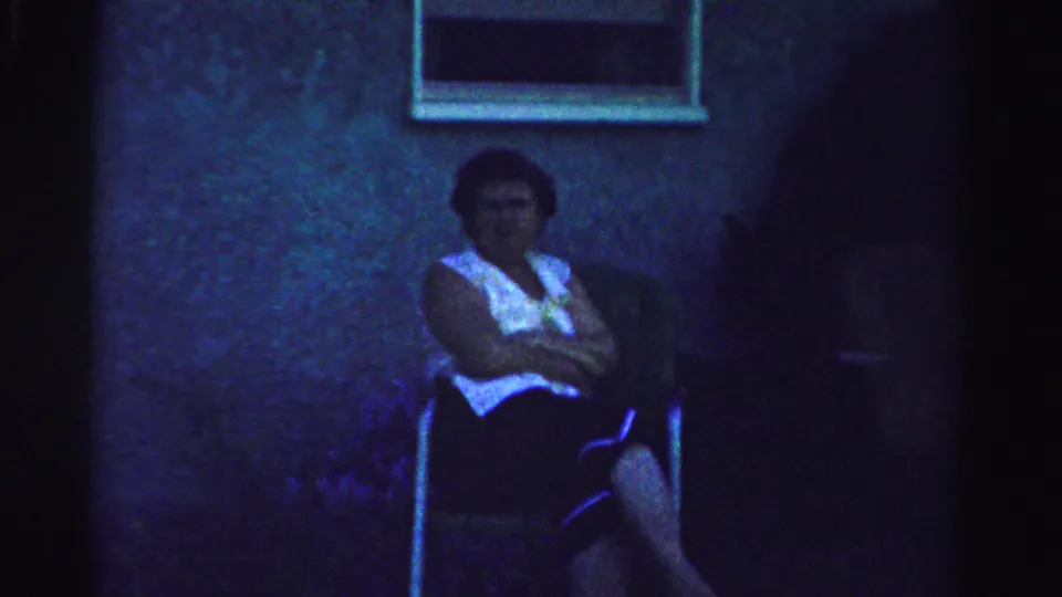 Older woman sits on a lawn chair by a house wall and window in low-light 1957 Normal, Illinois home-movie footage.