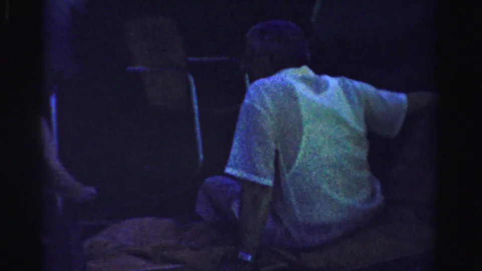 Older man in a light shirt sits close to camera while another seated figure is partly visible in shadow outdoors.