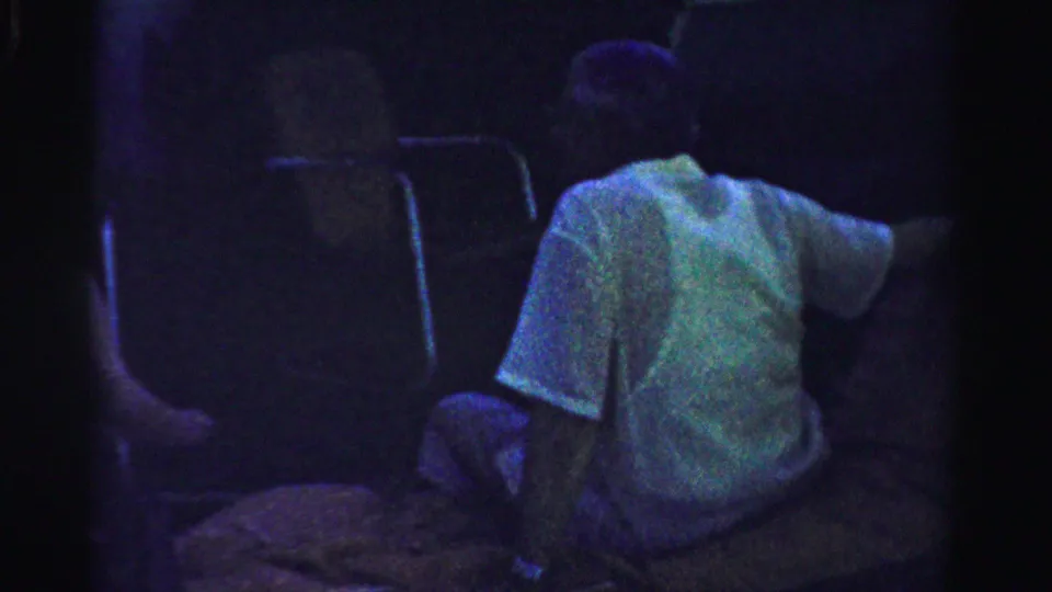 A man in a light shirt sits with his back partly turned in a dim outdoor setting in 1957 Normal, Illinois home-movie footage.