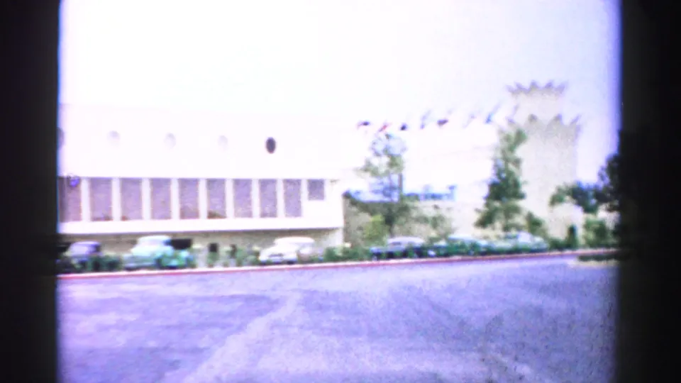 1957 Normal, Illinois 8mm frame of a large light-colored building with small flags, young trees, parked cars, and a broad paved drive.