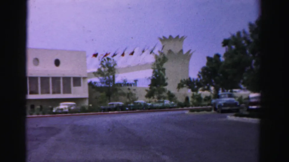 1957 Normal, Illinois 8mm frame showing the building exterior, rows of small flags, trees, mid-century parked cars, and a dark car at right.