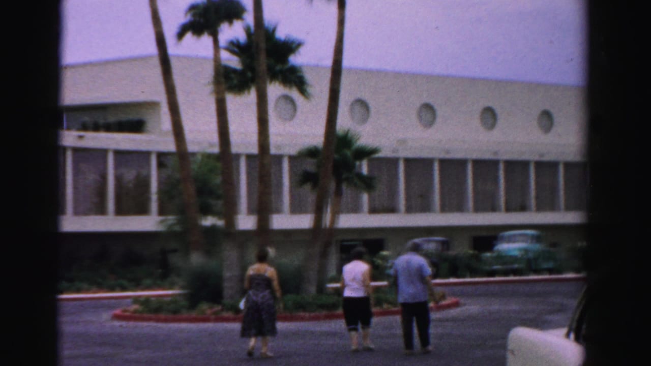 Adults walk across a hotel parking area near palms and parked 1950s cars.