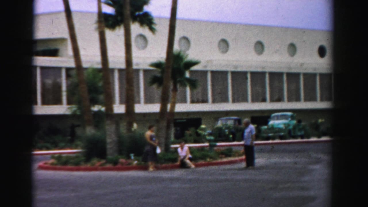 Adults stand and sit near a landscaped curb outside a modern hotel building.