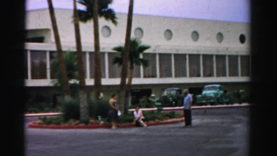 1957 Las Vegas frame with adults near hotel grounds, palms, parked cars, and a white facade with circular windows.