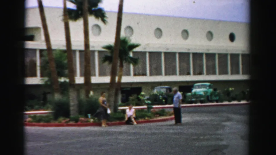1957 Las Vegas home-movie view of visitors by landscaped hotel grounds, with palms, round windows, and 1950s cars behind them.