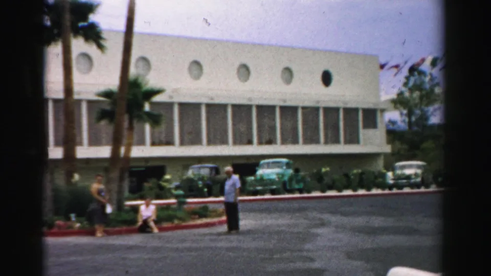 1957 Las Vegas frame showing a wider hotel exterior with visitors near the curb, palms, parked automobiles, and circular upper windows.
