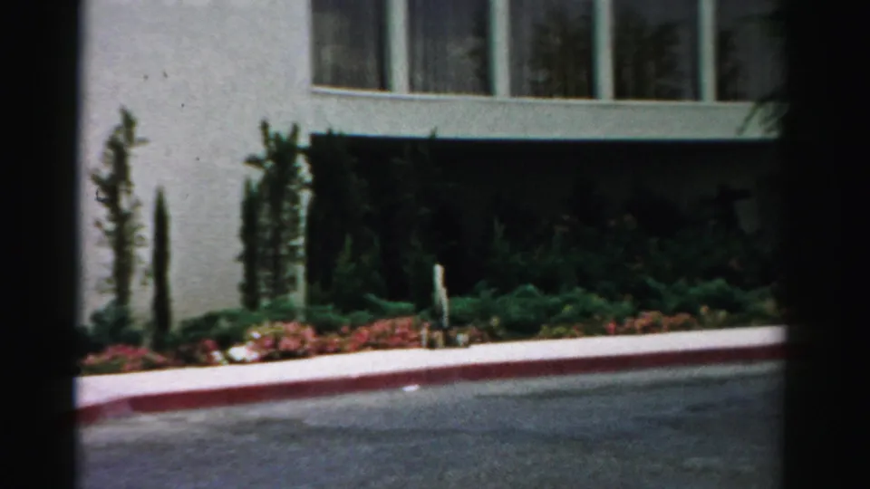 1957 Las Vegas exterior view with large upper windows, a dark overhang, dense garden plantings, sidewalk, red curb, and street pavement.