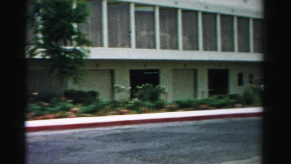 1957 Las Vegas frame showing the lower facade of a modern building with shrubs, dark door openings, a red curb, and asphalt.