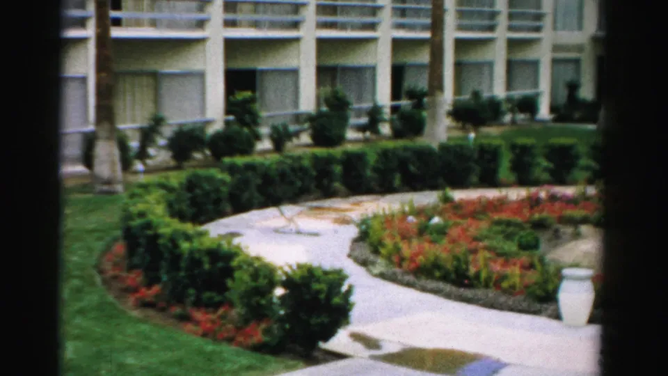 1957 Las Vegas 8mm frame of a landscaped garden path, lawn, shrubs, flowers, and a columned building exterior.