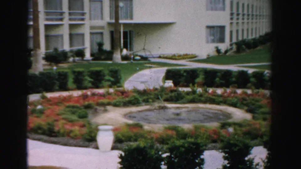 1957 Las Vegas apartment complex garden with a circular fountain, red flower beds, low hedges, pale walkways, lawn, and part of the building entrance.