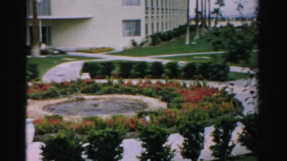 1957 Las Vegas apartment grounds with a circular fountain, red-orange garden beds, clipped hedges, pale paths, lawn, palm trunks, and the building facade.