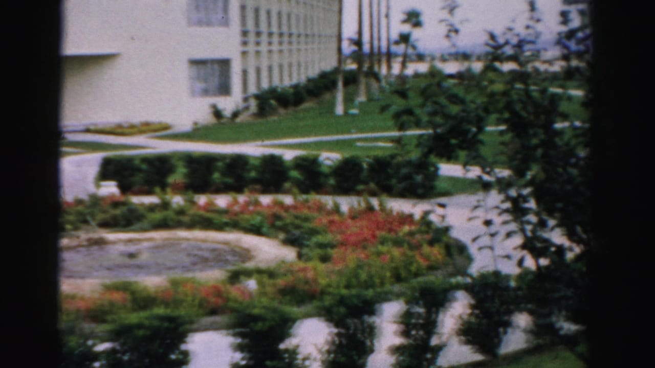 Manicured resort garden with flower beds, lawn, walkways, fountain edge, and light-colored buildings.