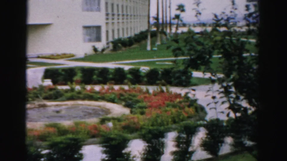 1957 Las Vegas resort garden view with a circular fountain edge, planted beds, clipped lawn, paths, and pale buildings in soft 8mm color.