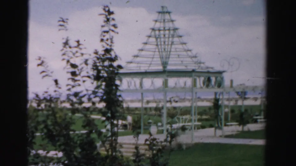 White trellis structure on 1957 Las Vegas resort grounds, with steps, lawn, shrubs, and soft 8mm exposure under a cloudy sky.