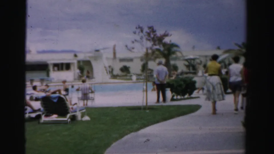 1957 Las Vegas resort pool area with guests walking on a curved path, people lounging near the water, lawn, chairs, and palm trees.