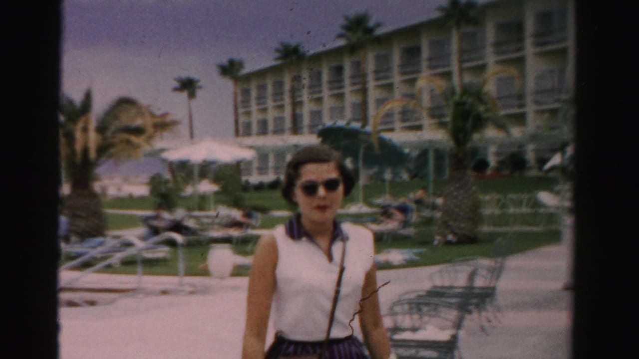 In 1957 Las Vegas vacation footage, a woman in a white sleeveless top and sunglasses walks toward the camera across hotel grounds with palm trees, lounge chairs, umbrellas, and a multi-story resort building behind her.