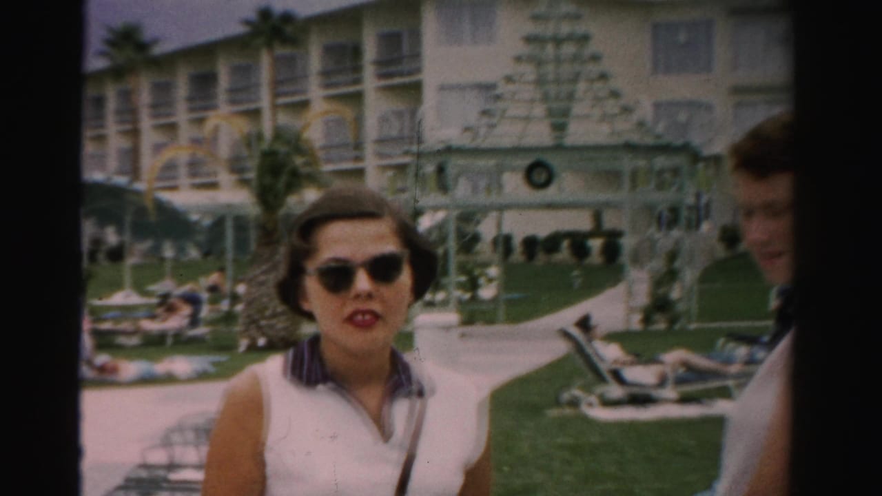 A woman in sunglasses approaches the camera in a Las Vegas hotel pool area, with resort balconies, palm trees, lawn, lounge chairs, and a partially visible adult man at the edge of the frame.