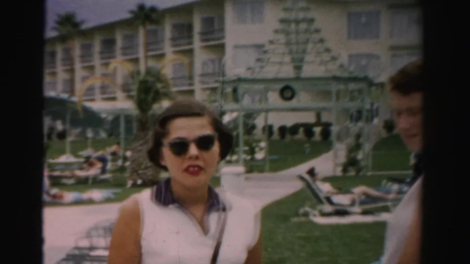A woman in sunglasses approaches the camera in a Las Vegas hotel pool area, with resort balconies, palm trees, lawn, lounge chairs, and a partially visible adult man at the edge of the frame.