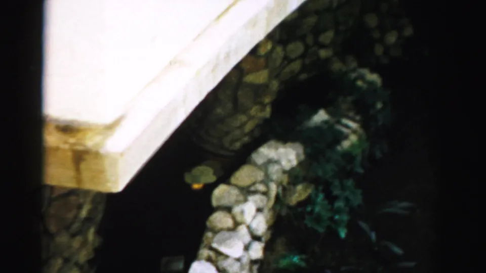 1957 Las Vegas 8mm frame showing a white exterior wall above stonework and greenery, with dark staining and soft home-movie exposure.
