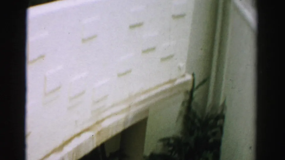 Close 1957 Las Vegas 8mm frame of a pale exterior wall with discoloration, shadowed seams, and a plant partly visible below.
