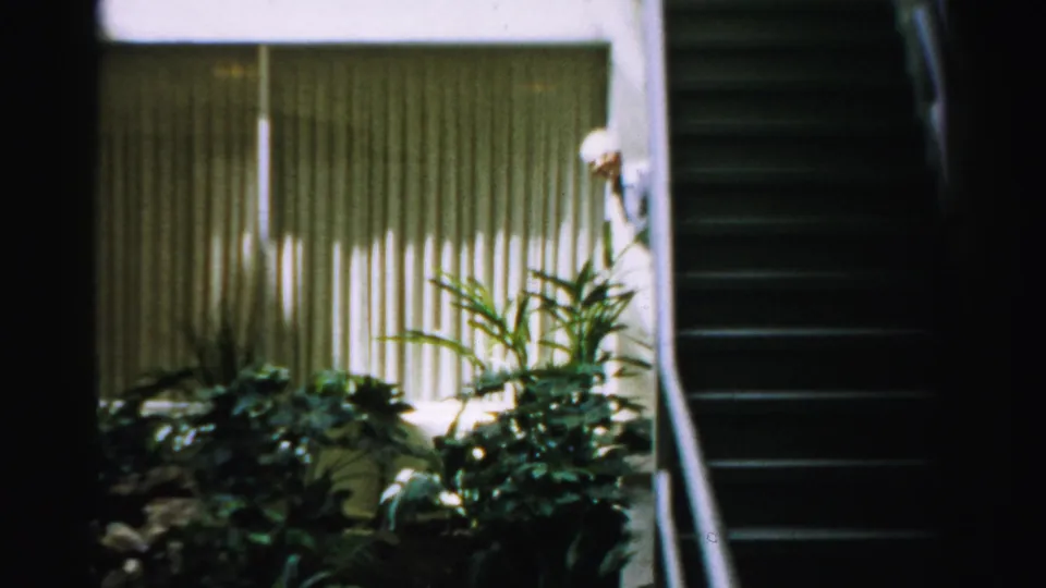 1957 Las Vegas home-movie frame showing vertical blinds, houseplants, a dark staircase, and a partly hidden person near the stairs.