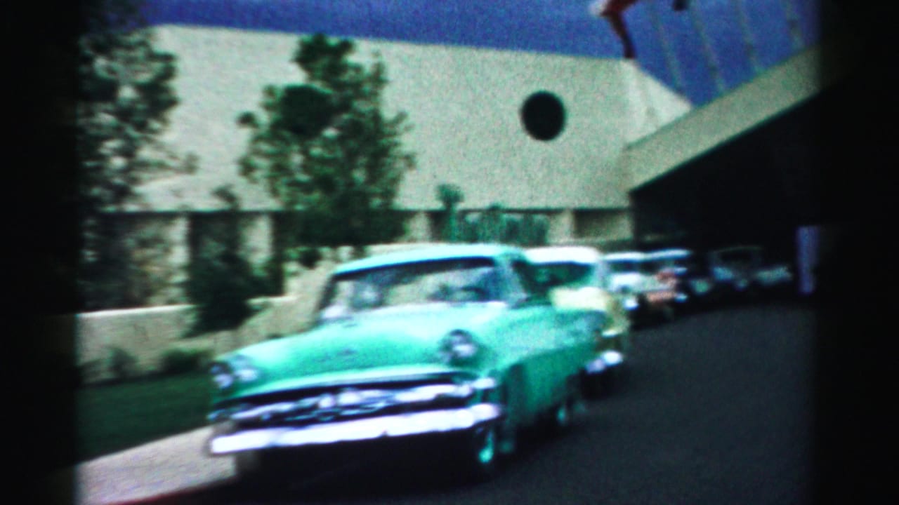 Turquoise 1950s car passing parked cars outside a pale hotel facade.