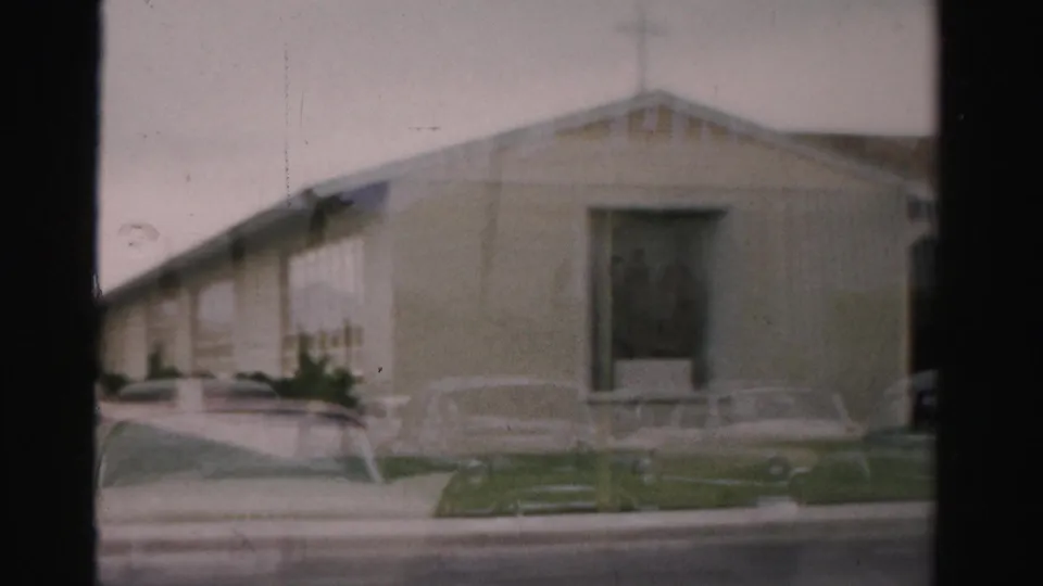 1957 Las Vegas frame showing the church front and side wall, a cross form near the roofline, parked cars, lawn, and cloudy sky.