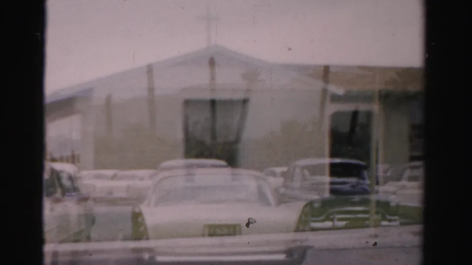 Soft 8mm frame of Las Vegas street frontage with parked 1950s cars and a pale low-rise building in the background.