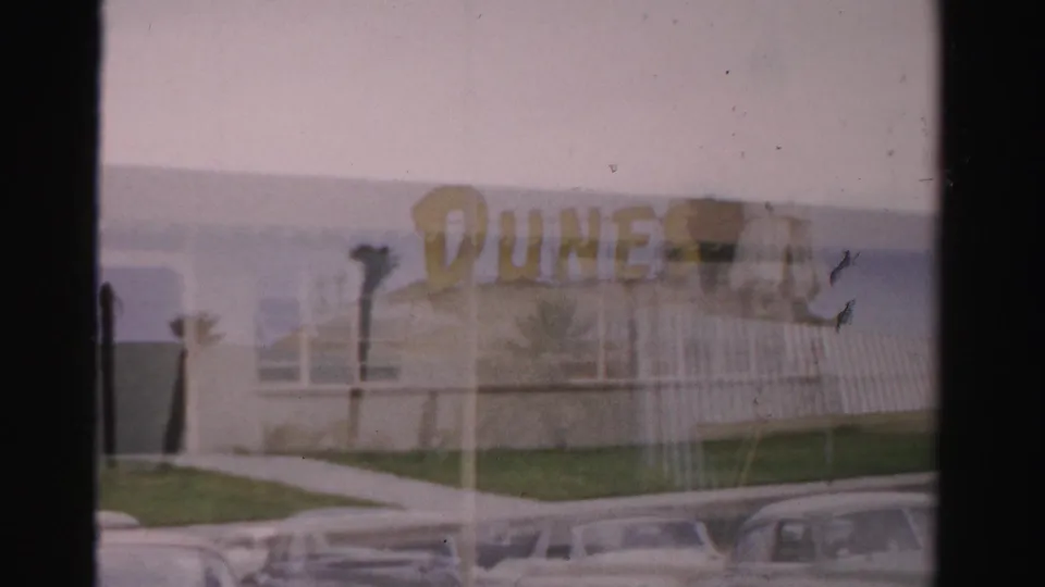 1957 Las Vegas view of a yellow DUNES sign on a low resort building, with lawn, palms, and parked cars visible.
