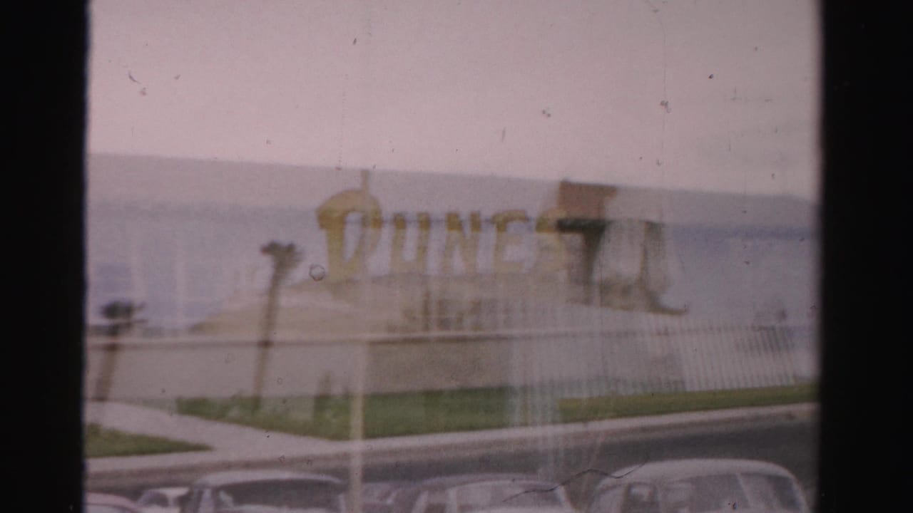 Soft 8mm frame of a school sign and parked mid-century cars near a low building.