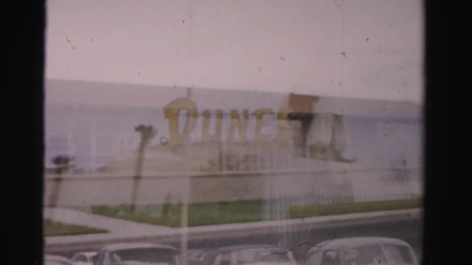 1957 Las Vegas home-movie frame showing part of the Gorman High School sign, a low facade, and parked mid-century cars.