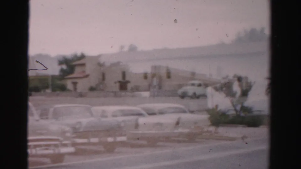 1957 Las Vegas home-movie frame showing mid-century cars in a parking lot with palms and low resort-style buildings beyond.