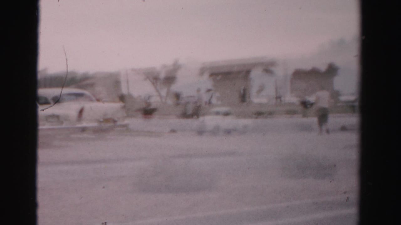 Paved lot with a parked car, low buildings, palms, and a distant person.