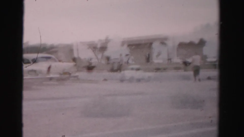 1957 Las Vegas frame with a parked car, palms, low building fronts, and a person walking across the lot.