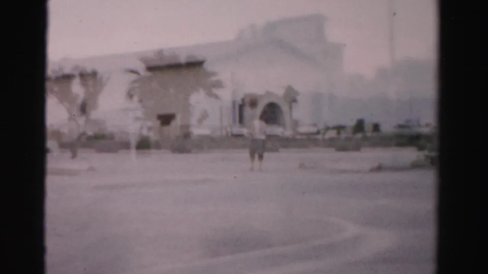 Soft 1957 Las Vegas 8mm frame showing a person near an arched low building, palms, and an open parking area.