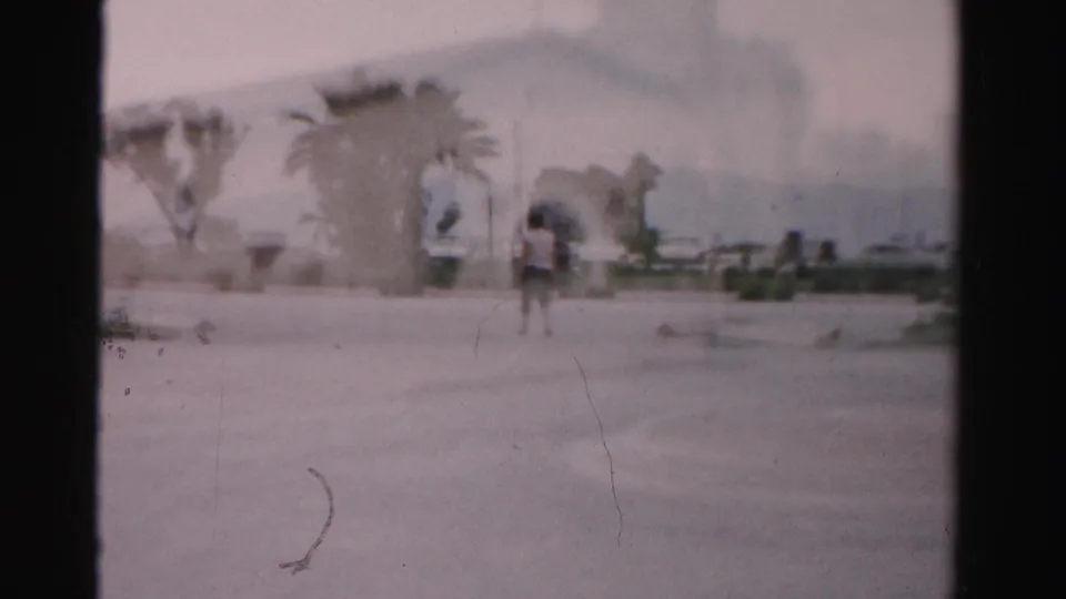 1957 Las Vegas home-movie frame with a distant person in a paved lot, palms, low buildings, and parked cars beyond.