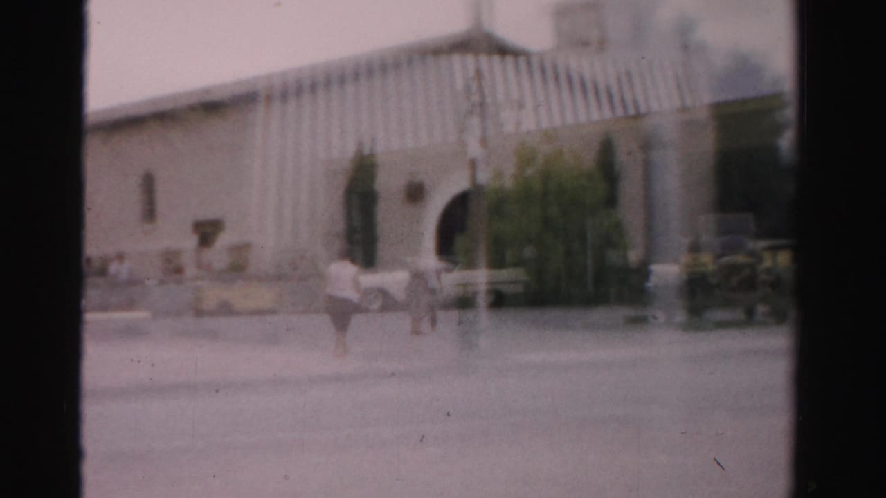 Pedestrians cross a hazy street in front of an arched stucco building and parked cars.