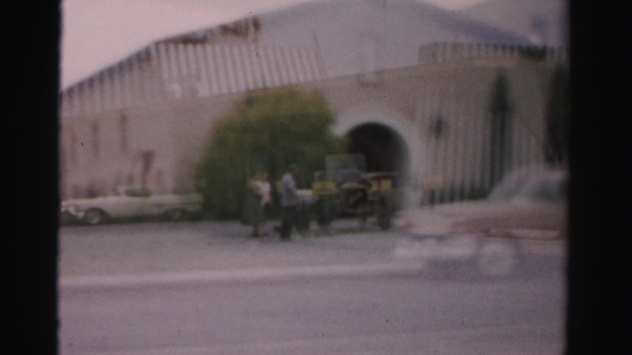 People stand or walk near parked vintage cars outside a large arched building.