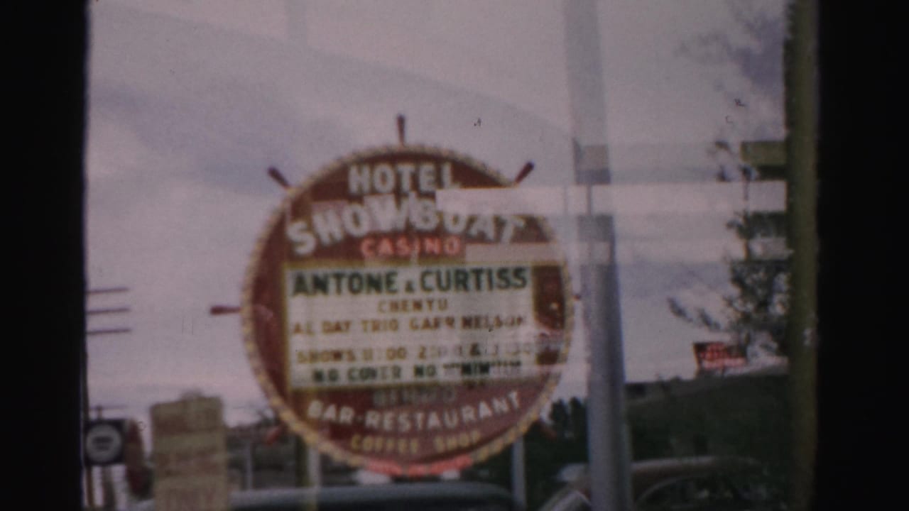 1957 Las Vegas view through glass of the Hotel Showboat Casino sign advertising live acts, with street poles and cars in the background.